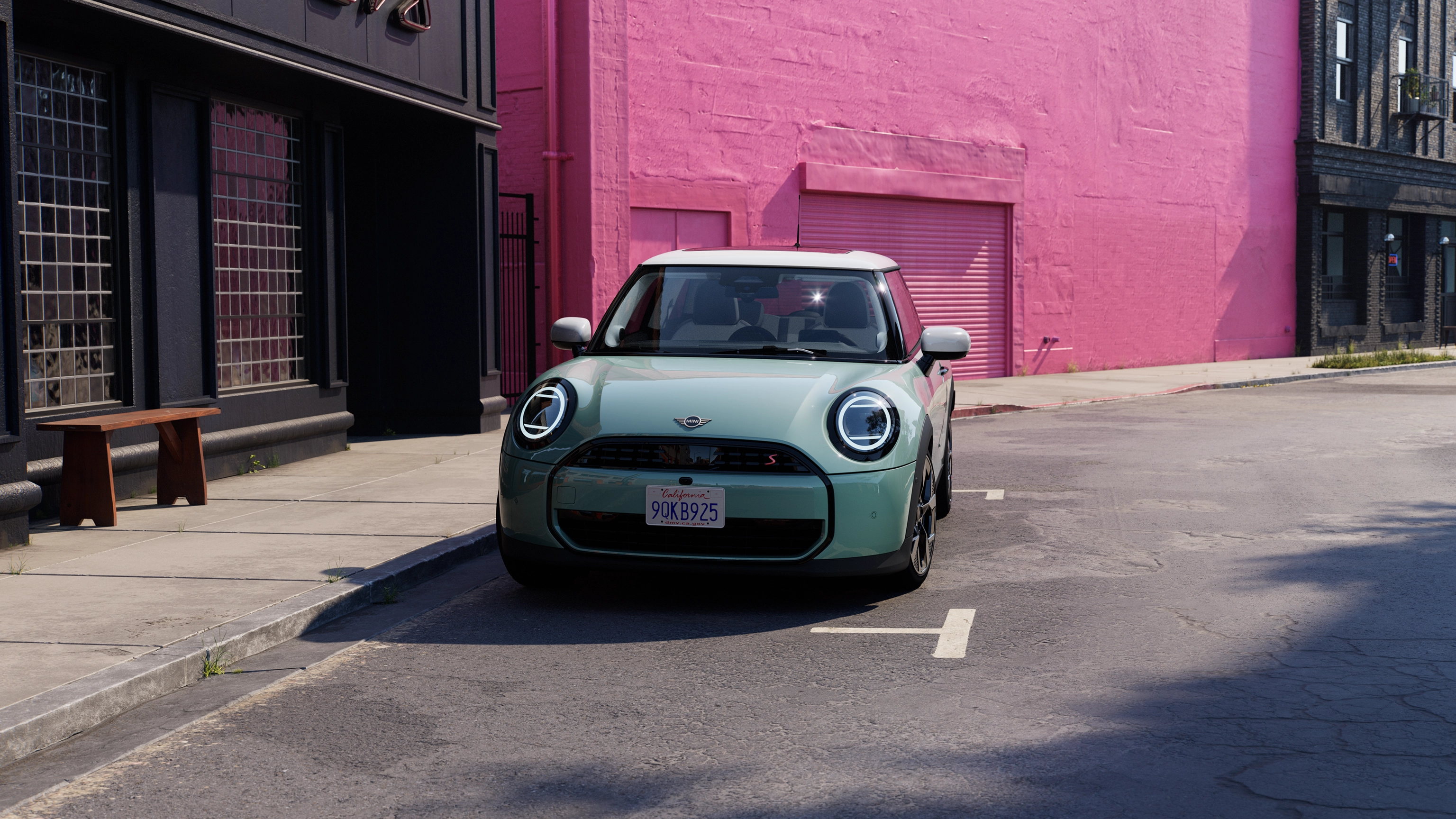 Frontansicht eines parkenden MINI Cooper in Ocean Wave Green mit Dach in Weiß auf einer Straße mit einer rosafarbenen Wand im Hintergrund