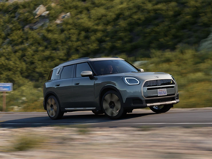 Seitenansicht eines im Gelände fahrenden vollelektrischen MINI Countryman in Smokey Green mit Dach in Vibrant Silver.