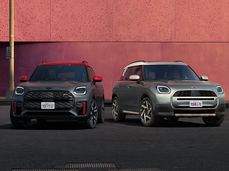Frontansicht des MINI Countryman JCW in Legend Grey mit rotem Dach und des MINI Countryman in Smokey Green mit Dach in Vibrant Silver, die nebeneinander stehen.