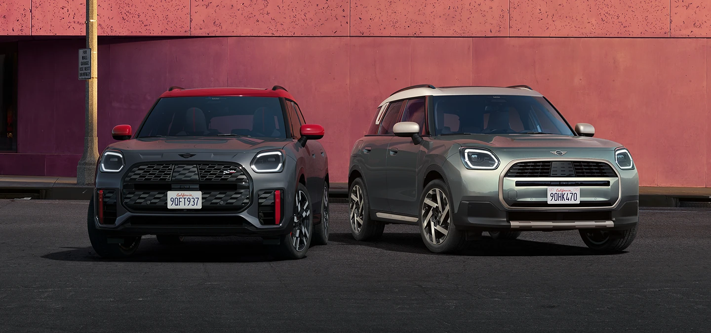 Frontansicht des MINI Countryman JCW in Legend Grey mit rotem Dach und des MINI Countryman in Smokey Green mit Dach in Vibrant Silver, die nebeneinander stehen.