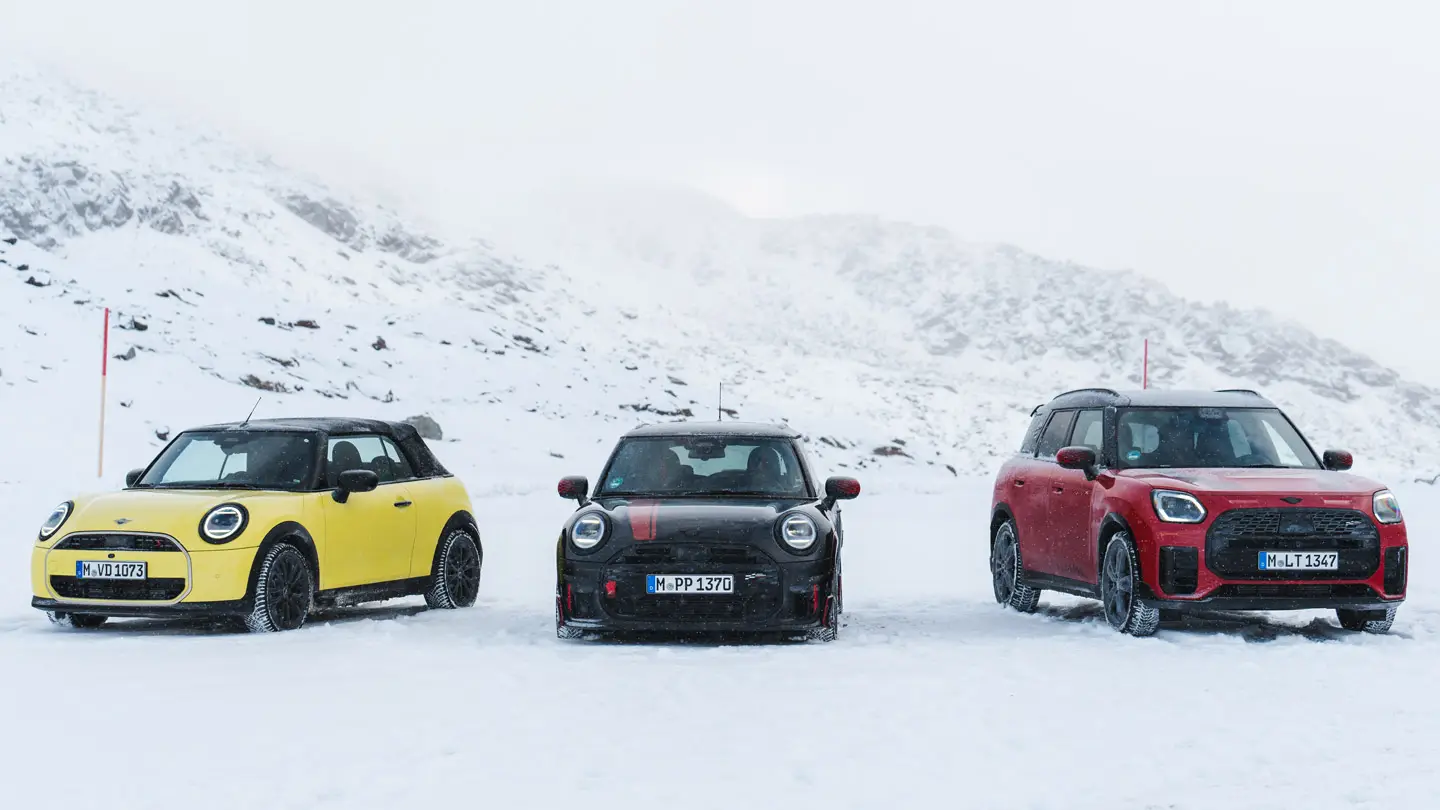 Ein MINI Cooper Cabrio in Sunny Side Yellow, ein MINI John Cooper Works in Midnight Black II und ein MINI Countryman in Chili Red stehen auf einer verschneiten Bergstraße in der Frontansicht.