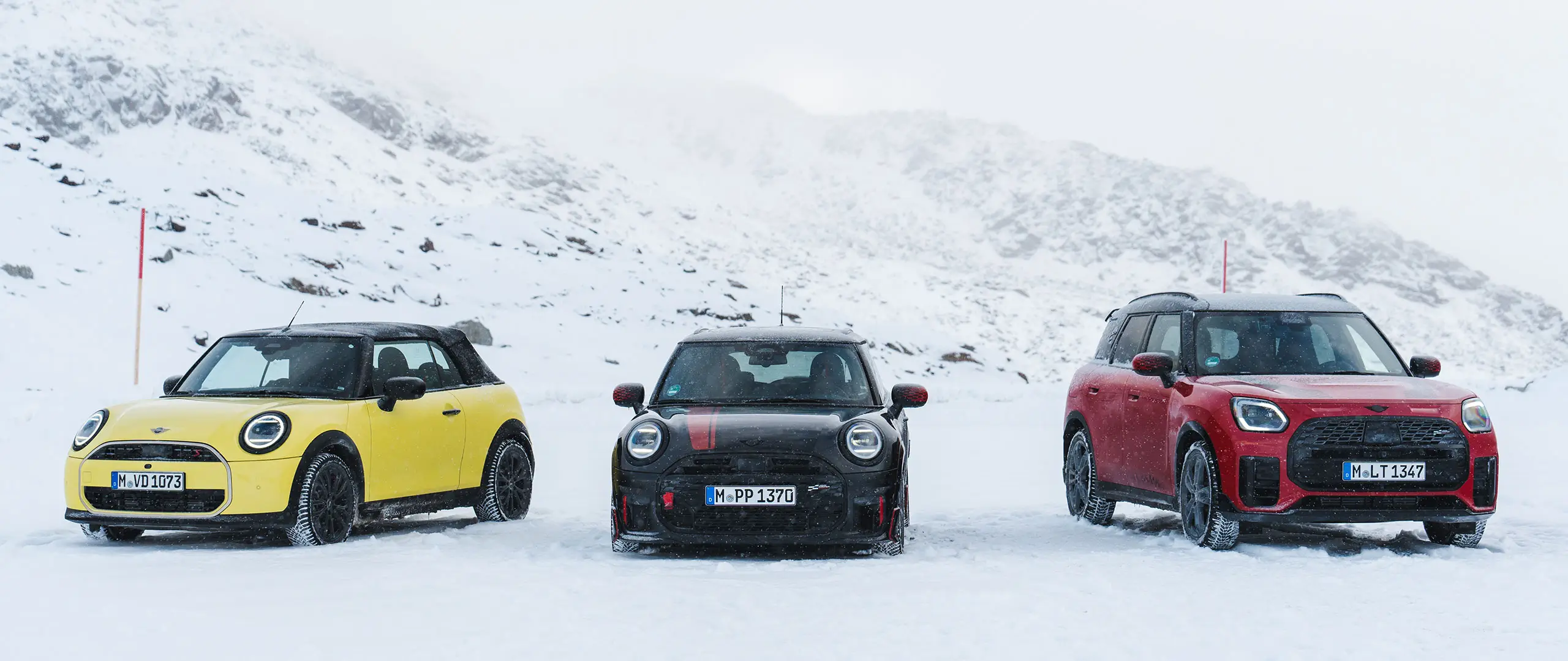 Ein MINI Cooper Cabrio in Sunny Side Yellow, ein MINI John Cooper Works in Midnight Black II und ein MINI Countryman in Chili Red stehen auf einer verschneiten Bergstraße in der Frontansicht.