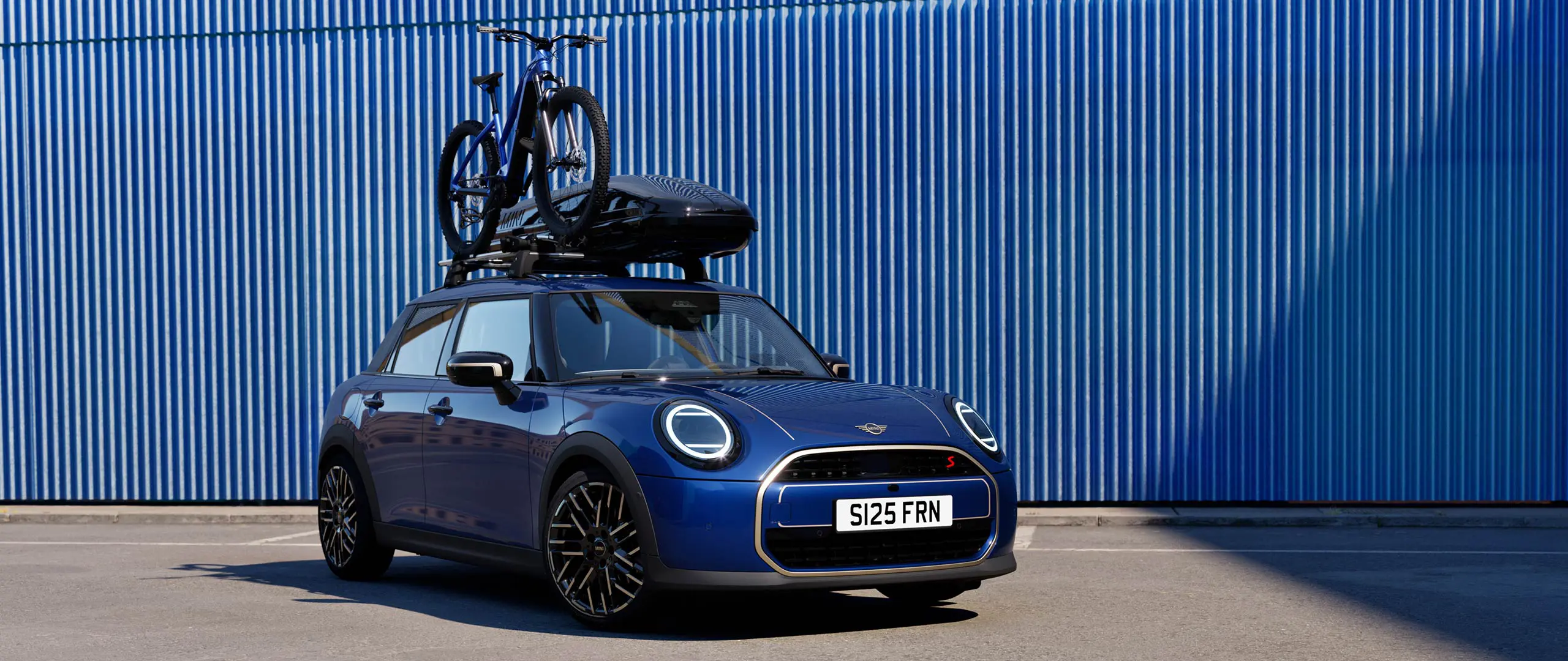 Vorderansicht des MINI Aceman in Indigo Sunset Blue mit MINI Fahrraddachträger mit Mountainbike und MINI Dachbox 320.