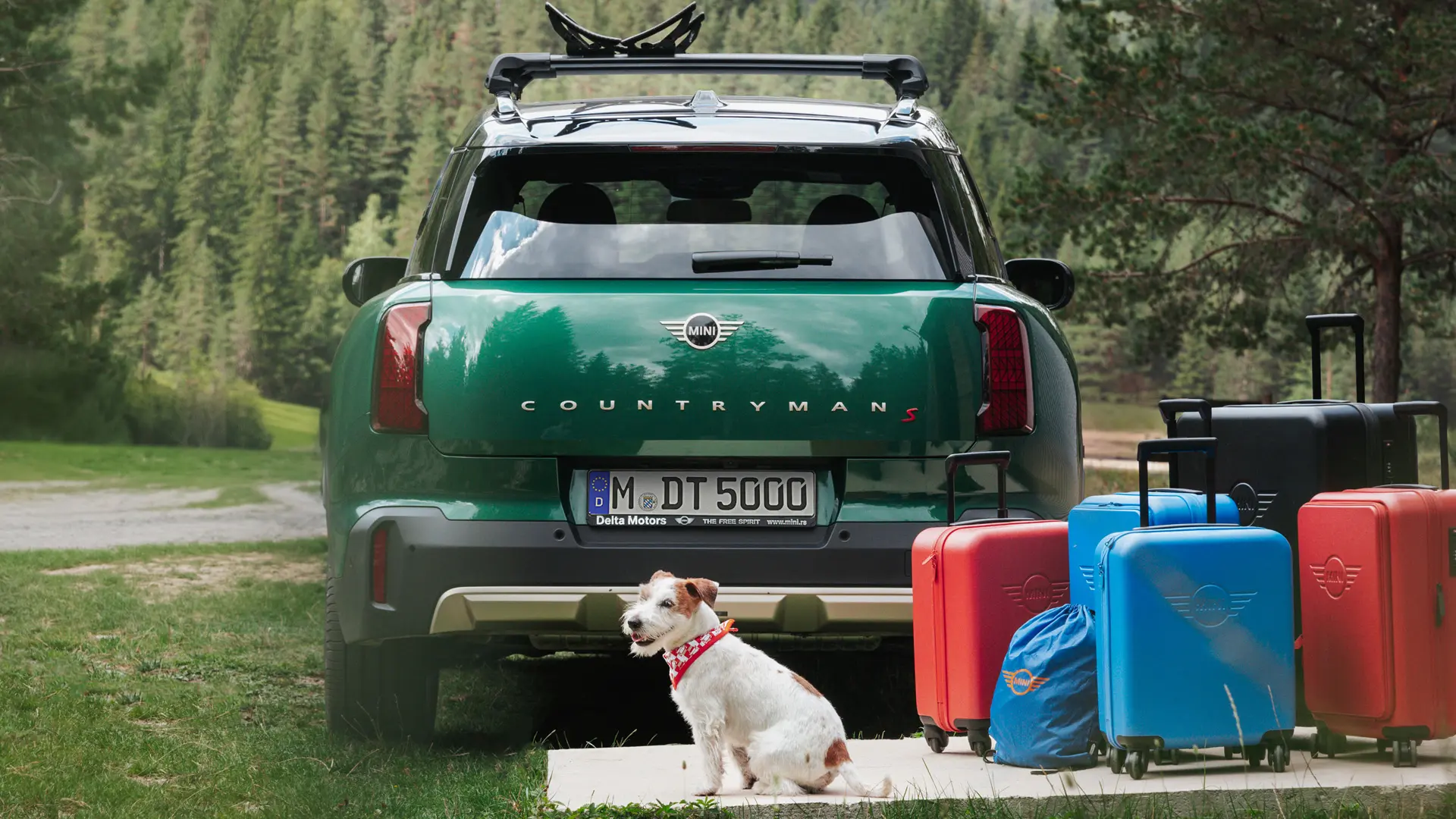 MINI Countryman in grün mit verschiedenen Gepäckstücken aus der MINI Lifestyle Collection, die zusammen mit einem Jack Russell Hund hinter dem Fahrzeug stehen. 