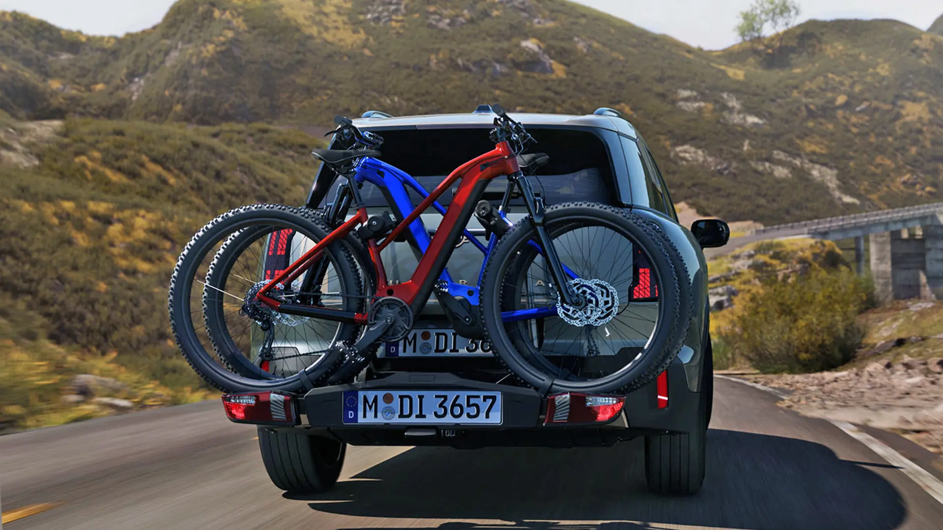 MINI Countryman in Smokey Green mit Fahrradheckträger Pro 3.0 und zwei Mountainbikes, Rückansicht, Fahrt auf einer Bergstraße.