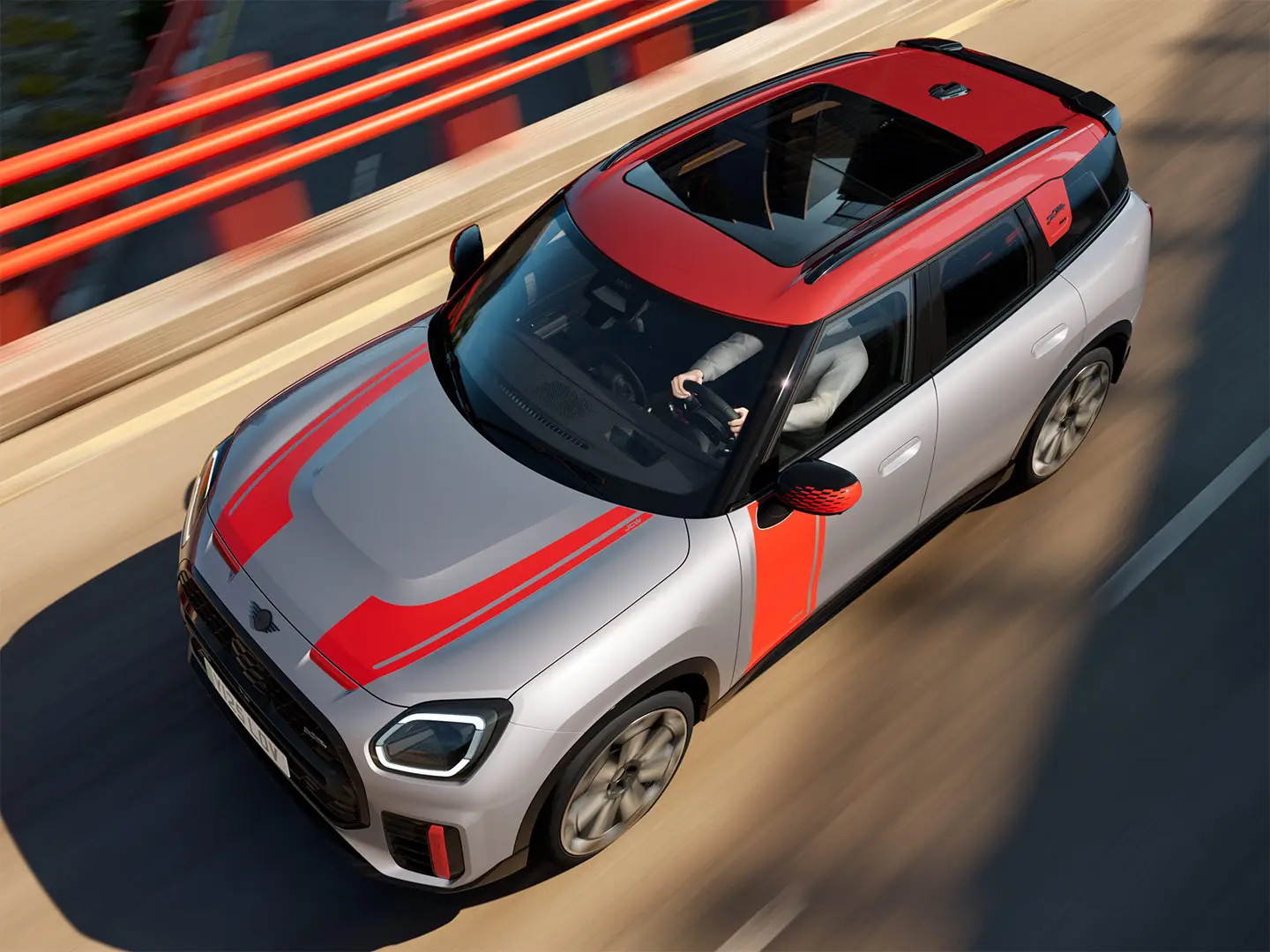 Ein MINI Countryman John Cooper Works in Melting Silver mit Spiegelkappe, Dekoraufklebern an Seite und Motorhaube, Dach und Winglet in Chili Red fährt auf einer Straße in der Stadt – seitliche Frontansicht.