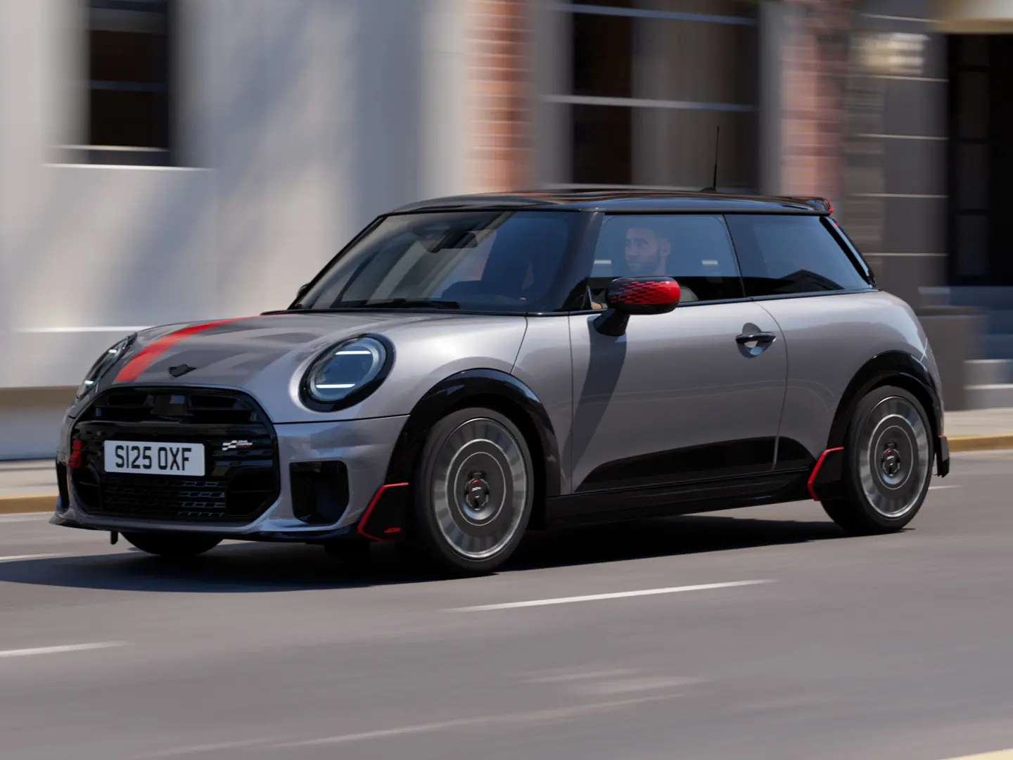 Ein MINI Cooper 3-Türer in Melting Silver mit John Cooper Works Spiegelkappe und Dekoraufklebern auf der Motorhaube in Chili Red, Dachkantenspoiler und Winglet in Schwarz fährt auf einer Straße in der Stadt – seitliche Frontansicht.