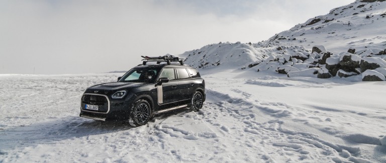 MINI Countryman in Silver auf einer Bergstraße mit MINI Dachträger, MINI Dachbox, MINI Fahrraddachträger mit montiertem Fahrrad und MINI John Cooper Works Winglet in der Frontansicht. 
