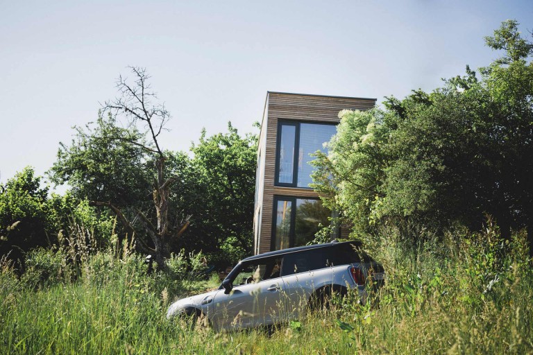 Motiv eines silbernen MINI Cooper SE, das Fahrzeug parkt im Grünen vor Böhms mlab.design Modulhaus. 